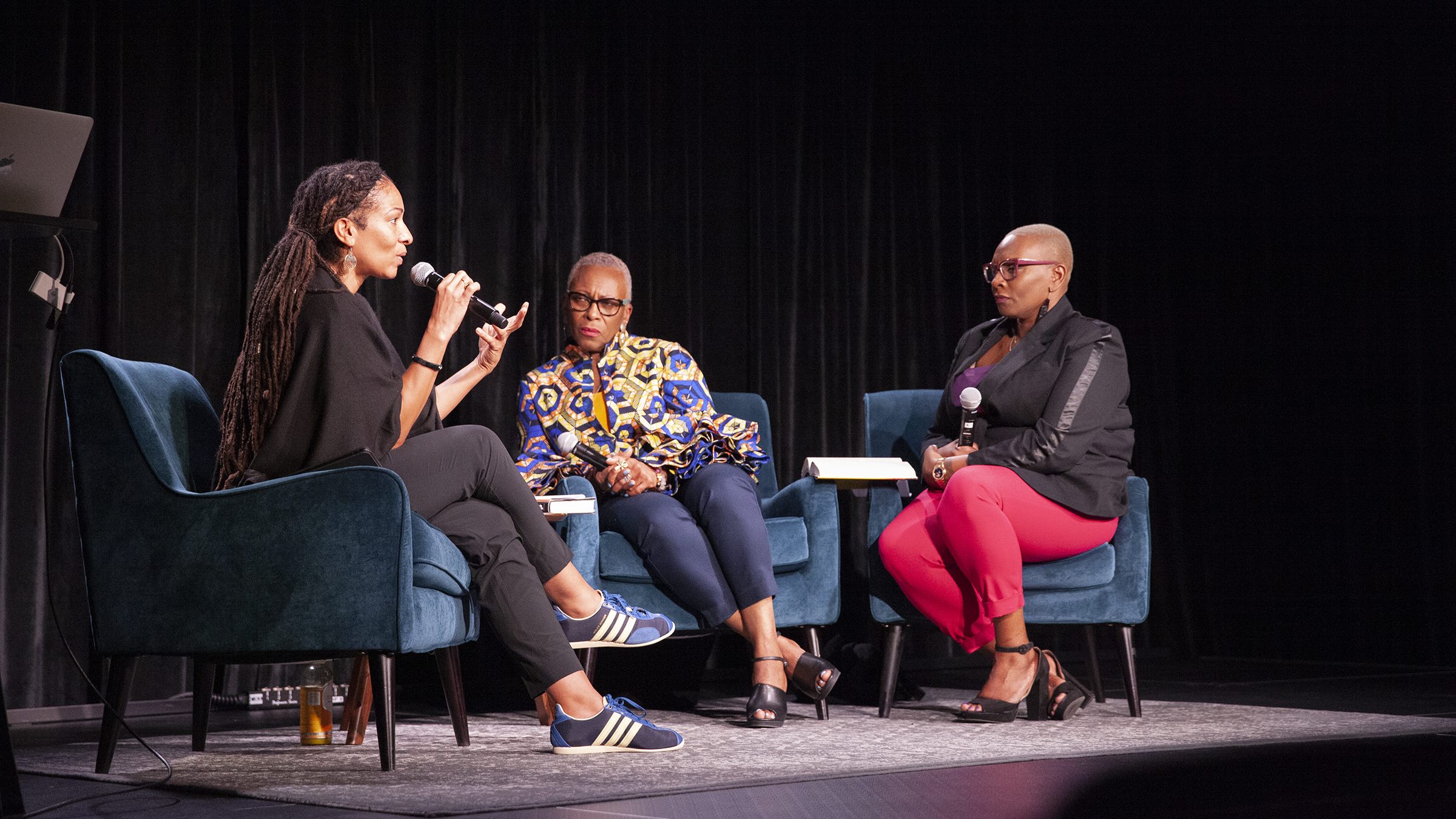 Three black women sitting on chairs on a stage, one holding a microphone